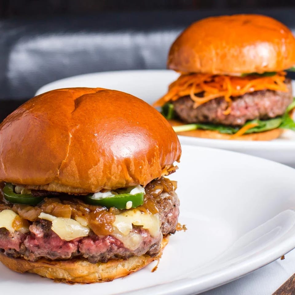 Two gourmet burgers on white plates, one with jalapeños and caramelized onions, the other with shredded carrots.