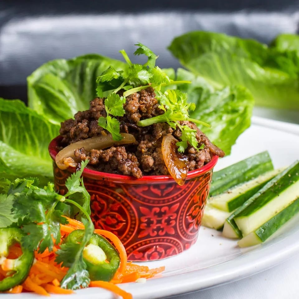 A red bowl of ground beef with onions and cilantro, surrounded by lettuce, cucumbers, and peppers.