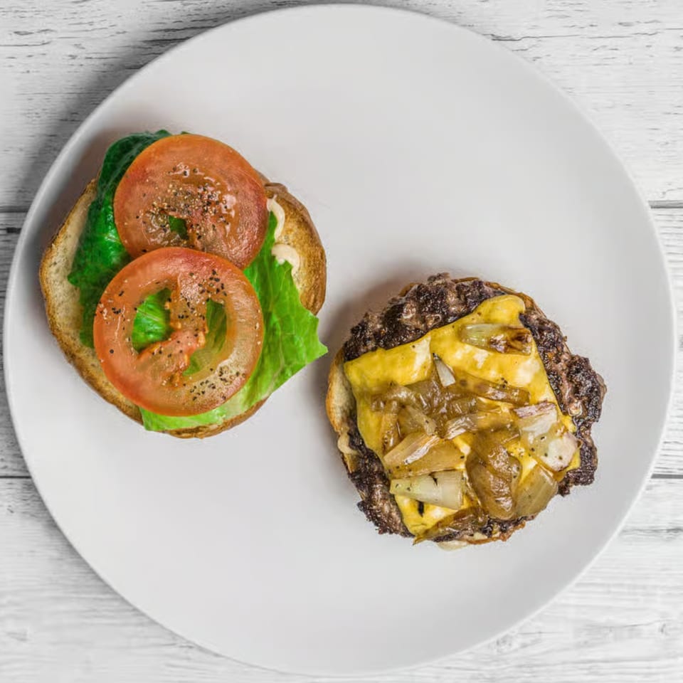 Deconstructed Burger Palace burger with cheesy patty, caramelized onions, lettuce, and tomato on a white plate.