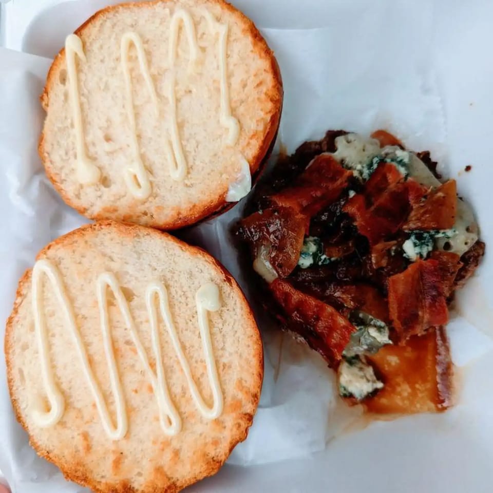 An open burger with toasted buns drizzled with sauce, bacon, and blue cheese crumbles.